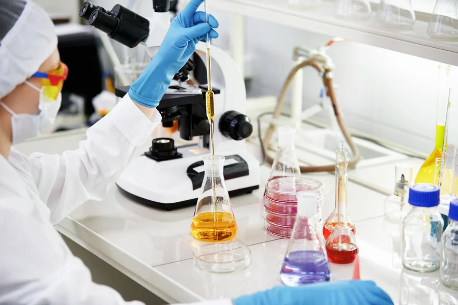 Analista de laboratorio pipeteando sobre un matraz, con microscopio y reactivos de colores en la mesa